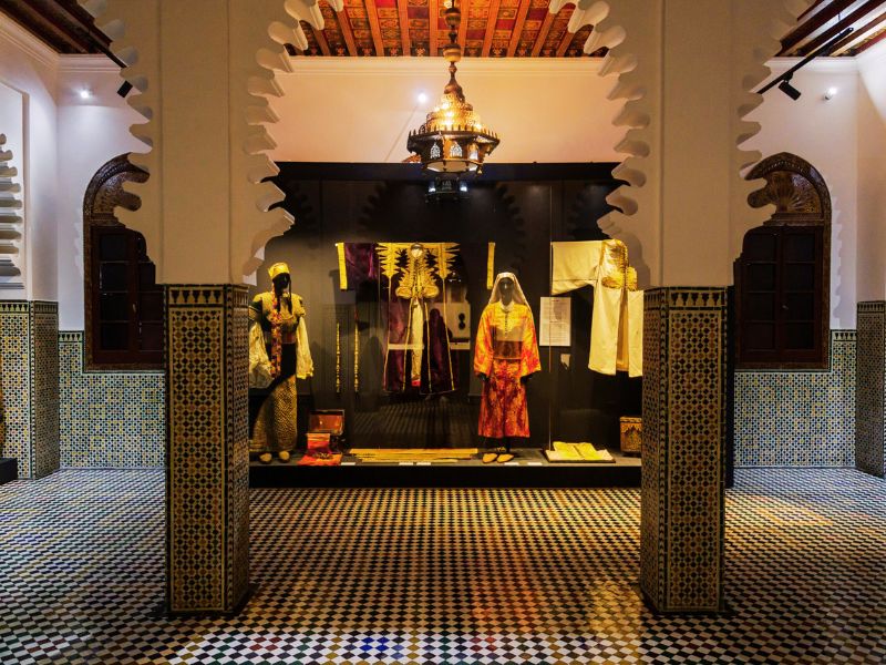 Interior view of the Ethnographic Museum of Tetouan, showcasing traditional costumes and artifacts against tiled walls and ornate architectural details.