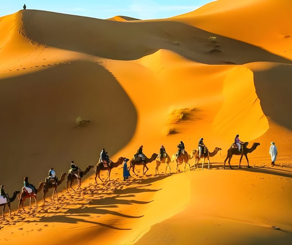 A caravan of camels and riders traversing golden sand dunes under a clear blue sky.