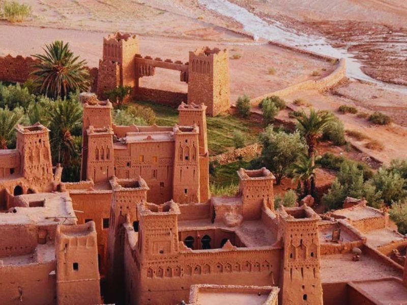 A scenic overview of Ksar Ait Benhaddou, showcasing its traditional earthen buildings and fortified structures amidst a backdrop of palm trees and arid landscape.