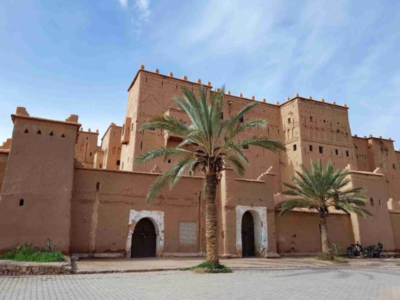 A view of a traditional Moroccan Kasbah featuring mud-brick architecture, palm trees in the foreground, and a blue sky.