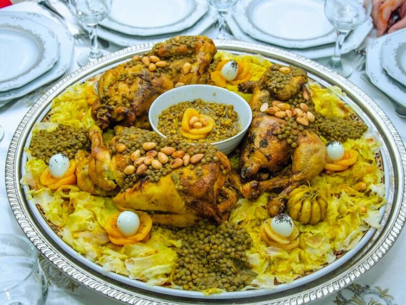 A beautifully arranged platter of Rfissa, featuring tender chicken pieces topped with lentils, almonds, and boiled eggs, served over a bed of shredded trid pancakes, with decorative orange slices.