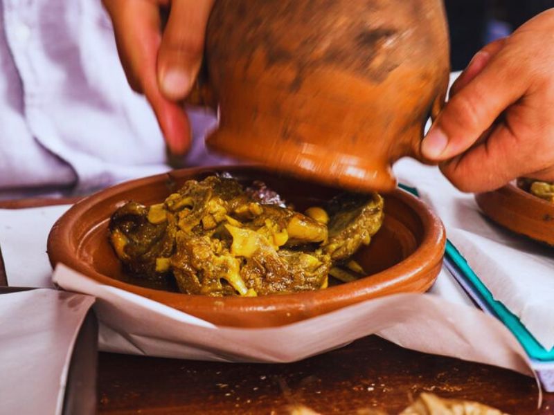 A person lifting a clay pot to reveal a traditional Moroccan dish, Tanjia, which contains succulent meat and spices in a terracotta bowl.