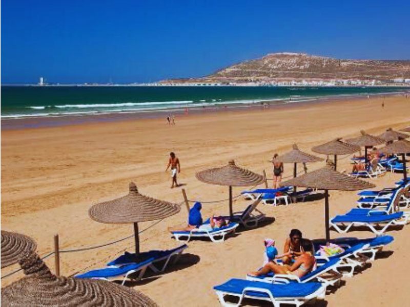 Agadir Beach in Morocco featuring golden sand, sun loungers, and beach umbrellas, with people enjoying the sun and the Atlantic Ocean in the background.