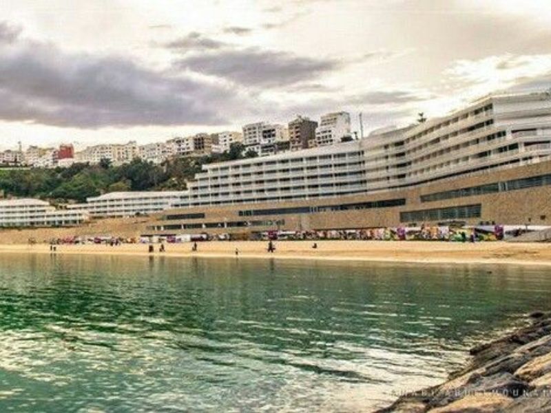View of Al Hoceima's beachfront with modern buildings overlooking the sea, featuring sandy shores and gentle waves under an overcast sky.