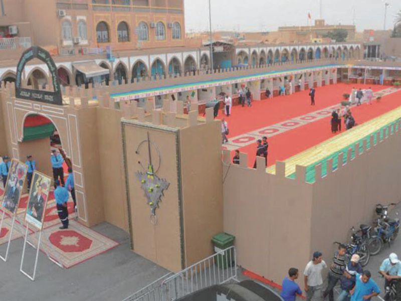 A view of the entrance to the Festival of Timizart in Tiznit, Morocco, featuring vibrant decorations and attendees walking around a red carpet area.