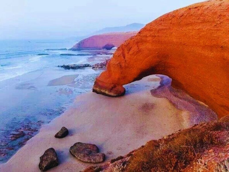 A view of Legzira Beach in Morocco, featuring a striking red sandstone arch, sandy shores, and gentle ocean waves in the background.