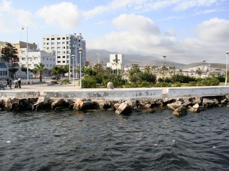 A view of a waterfront promenade in Nador, Morocco, featuring modern buildings, greenery, and the Mediterranean Sea in the foreground under a partly cloudy sky.