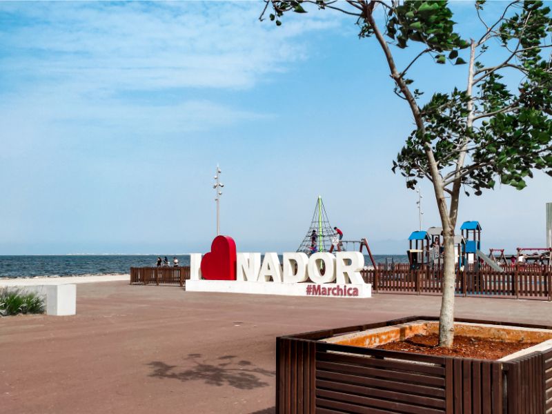 A public space in Nador featuring a large sign reading 'I ❤️ NADOR #Marchica' with playground equipment in the background, alongside the Mediterranean Sea under a blue sky.