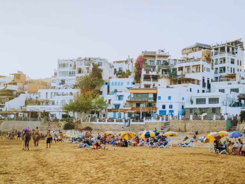 A scenic view of Taghazout Beach, featuring a sandy shoreline populated with sunbathers and colorful umbrellas, while buildings with white facades and greenery overlook the beach.
