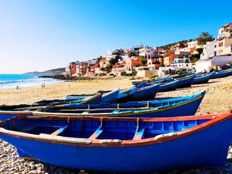 A scenic view of Taghazout Beach in Morocco, featuring traditional blue fishing boats on the sand, with colorful buildings and palm trees in the background.