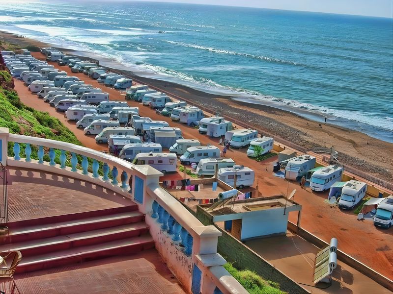 Aerial view of a coastline campsite in Sidi Ifni, Morocco, featuring several parked camper vans along a sandy beach with waves gently lapping at the shore.