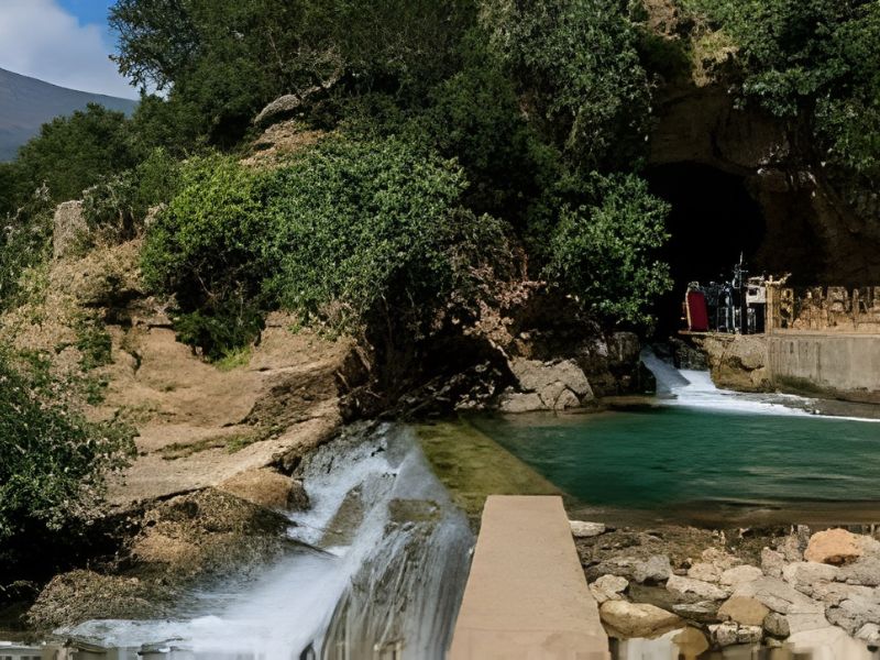A natural scene showcasing the Grotte des Pigeons in Tafoughalt, Morocco, featuring lush greenery, a clear water pool, and a small waterfall flowing into it.