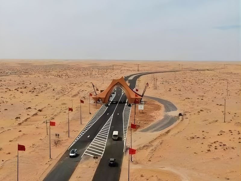 Aerial view of a modern gateway structure in Laâyoune, surrounded by sandy desert, with roads merging and Moroccan flags on display.