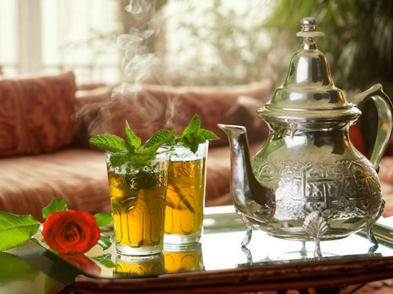 A silver Moroccan teapot with intricate designs next to two glasses of mint tea, garnished with fresh mint leaves, on a decorative tray. A rose and leaves are also visible, with a cozy interior setting in the background.