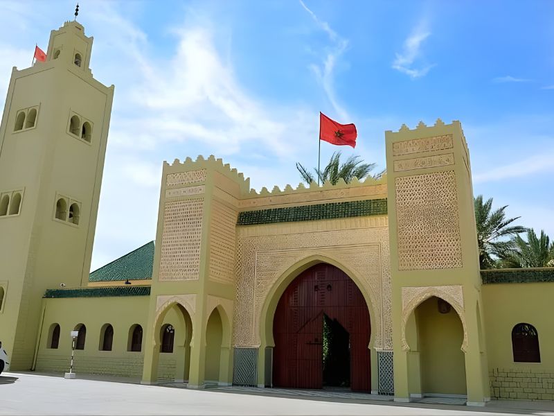 The entrance of a historical building in Rissani, Morocco, featuring traditional Moroccan architecture, a large arched doorway, and the Moroccan flag flying atop the structure.