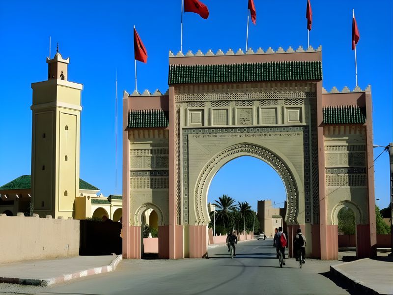A traditional architectural archway in Rissani, Morocco, with Moroccan flags adorning its top, welcoming visitors to the town.
