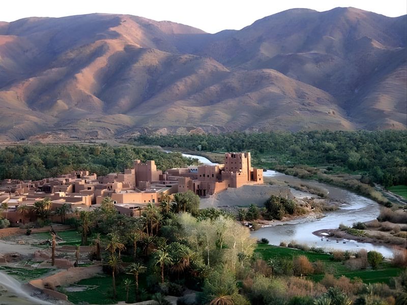 A scenic view of a Moroccan oasis with traditional adobe buildings nestled against a backdrop of mountainous terrain and lush greenery along a river.
