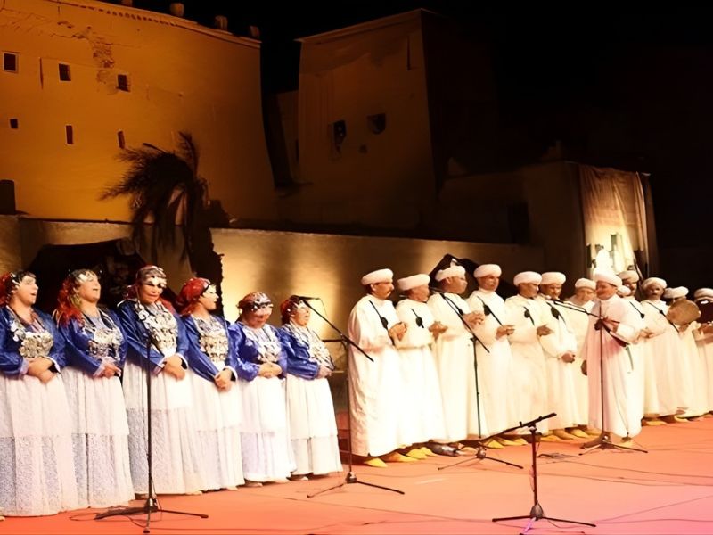 A group of Amazigh performers in traditional attire singing and dancing during an Ahwach celebration, with a backdrop of ancient architecture illuminated at night.