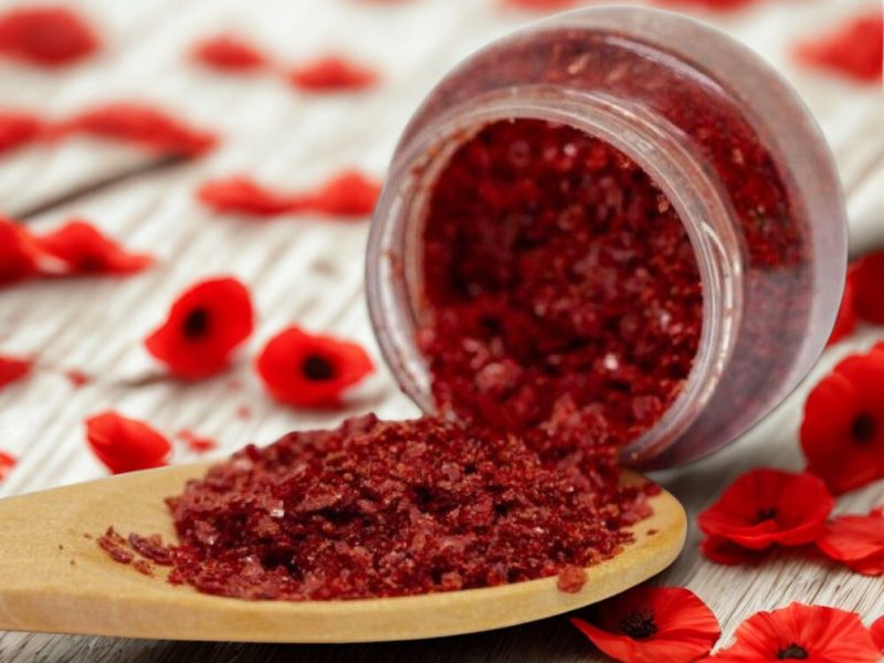 Aker Fassi, a deep red organic powder made from dried poppy petals and pomegranate rind, displayed in a wooden spoon beside a jar and surrounded by scattered poppy petals.
