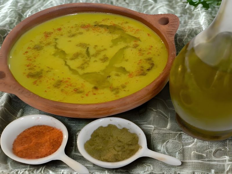 A bowl of Bissara, a Moroccan dried bean soup, topped with olive oil and spices, alongside small dishes of chili powder and cumin, with a bottle of olive oil in the background.