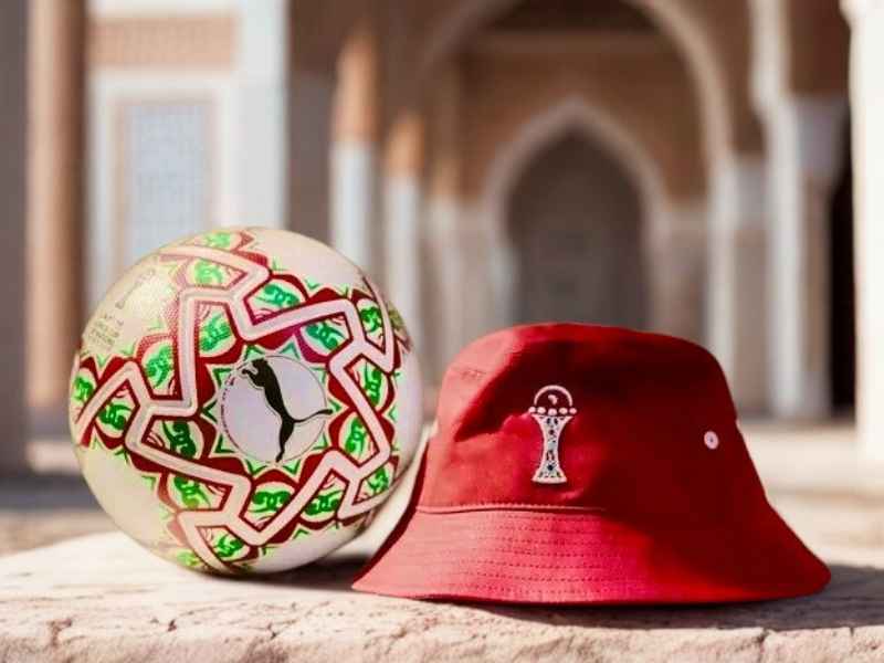 A soccer ball with a geometric design sits beside a red bucket hat featuring a logo, set against an ornate architectural background in Morocco.