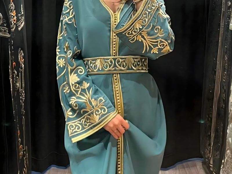 A person wearing a traditional Moroccan caftan with intricate gold embroidery, posing in front of a dark background.