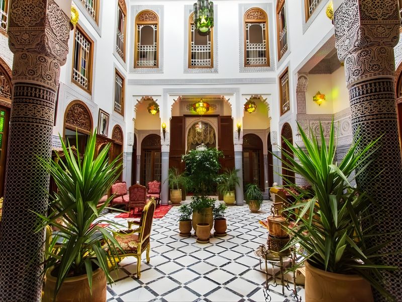 Interior view of a traditional Moroccan Riad featuring a courtyard with marble tiles, potted plants, and decorative arches.