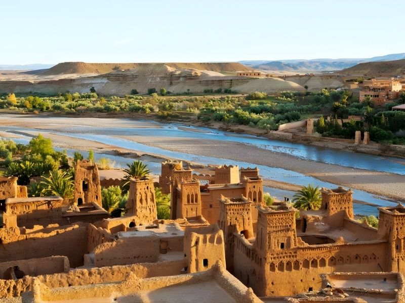 A scenic view of ancient earthen buildings surrounded by greenery and a river flowing through the landscape, with mountains in the background.