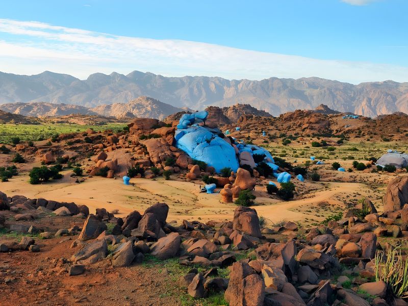 A landscape featuring rocky terrain with large boulders, some covered in blue material, and a backdrop of distant mountains under a clear blue sky.