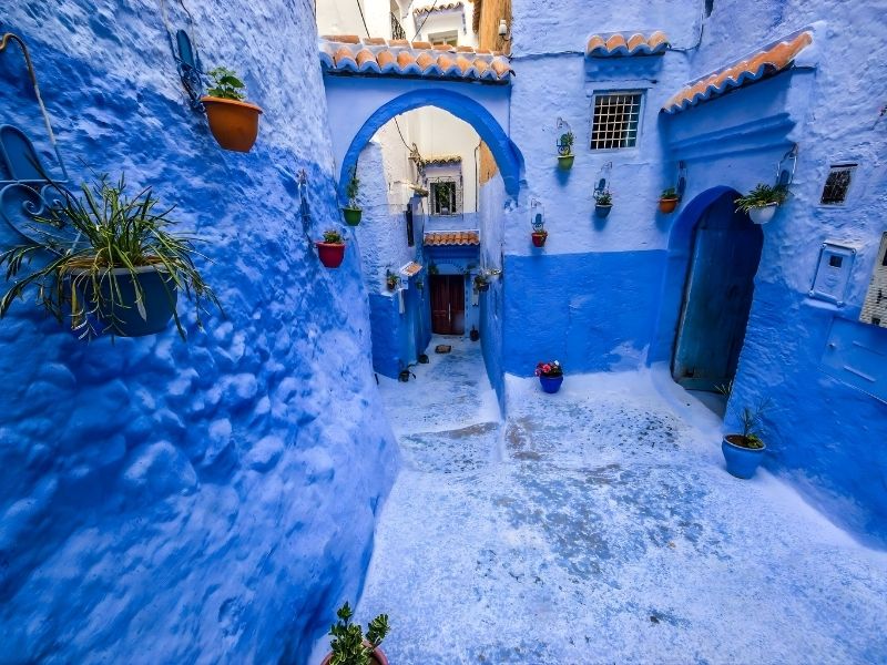 A picturesque alleyway in a blue-painted village, featuring decorative pots with plants, arched doorways, and a cobblestone floor.