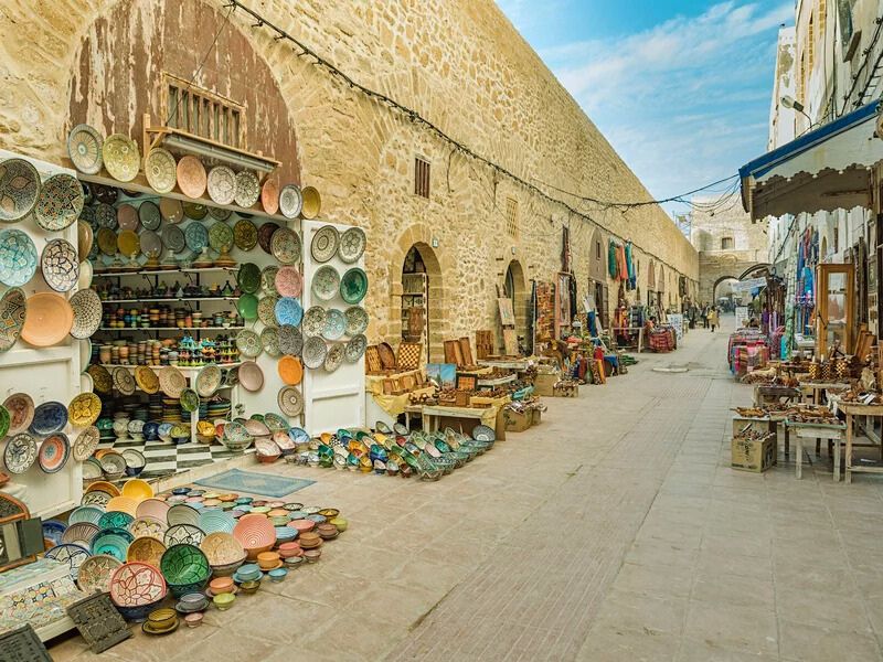 A narrow marketplace lined with stone walls, featuring an array of colorful pottery and handcrafted items displayed on the ground and on shelves. The scene showcases a vibrant atmosphere with various shops selling traditional crafts.