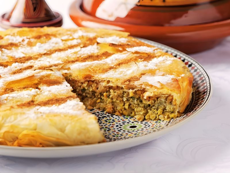 A beautifully presented Chicken Pastilla on a decorative plate, showcasing its flaky golden pastry crust and layered filling of shredded chicken, almonds, and spices. A portion has been cut out, revealing the intricate filling inside.