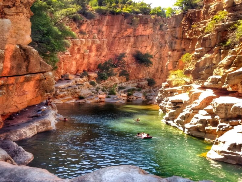 A scenic view of a natural pool surrounded by steep, rocky cliffs and lush greenery, with people swimming and kayaking in the water.