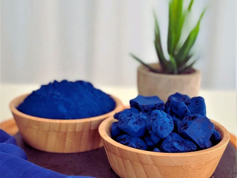 Two wooden bowls containing blue powder and blue chunks of material, with a small green plant in a pot in the background.