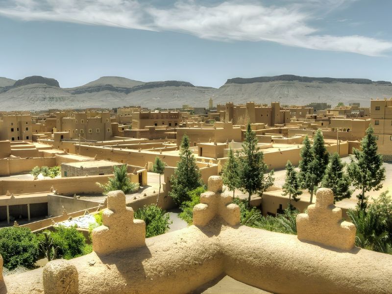 A panoramic view of a desert town with traditional clay architecture surrounded by mountains and greenery under a clear sky.