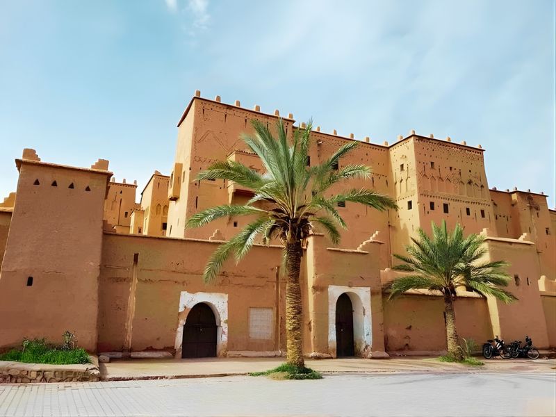 A view of a traditional adobe fortress with palm trees in the foreground under a blue sky.