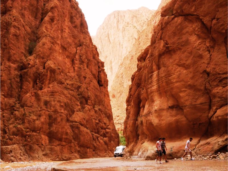 A narrow canyon with towering red rock walls, featuring a van parked on a rocky path and people walking through the canyon.