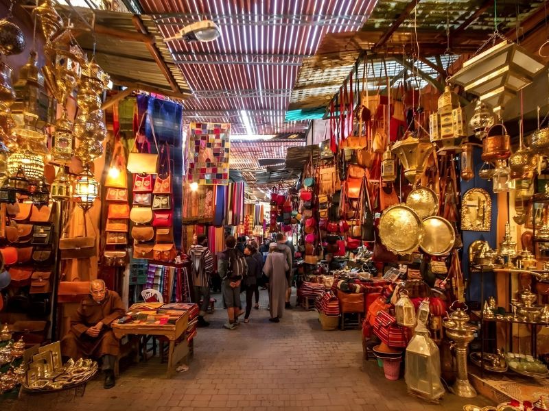 A bustling market street lined with colorful stalls selling handcrafted goods, textiles, and ornate lanterns, with people browsing and a person sitting at a table.