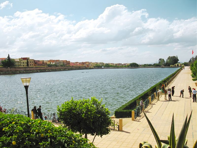 A scenic view of a river with buildings along the banks, surrounded by lush greenery and a paved walkway. People are seen enjoying the area under a partly cloudy sky.