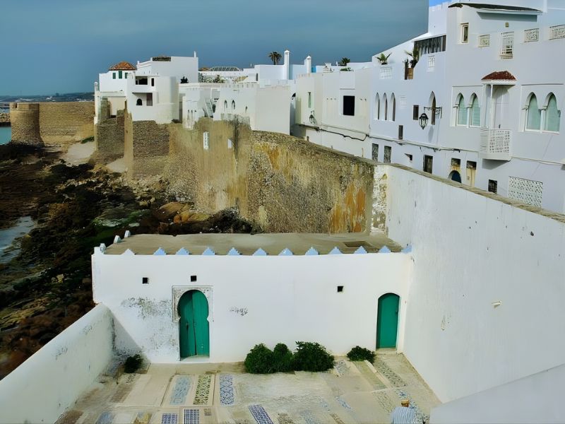 A coastal view of white buildings with green doors and windows, featuring a stone wall and rocky shoreline in the foreground.