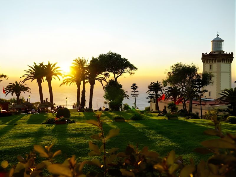 Scenic view of a sunset over the ocean, with palm trees and a lighthouse in the foreground, and manicured lawns illuminated by warm light.