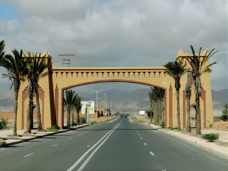 A large decorative archway with palm trees on both sides, standing at the beginning of a road with mountains in the background and cloudy skies above.