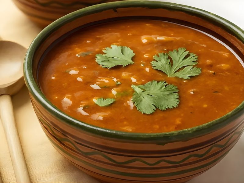 A bowl of rich, orange soup garnished with fresh cilantro leaves, placed on a light-colored tablecloth alongside a wooden spoon.