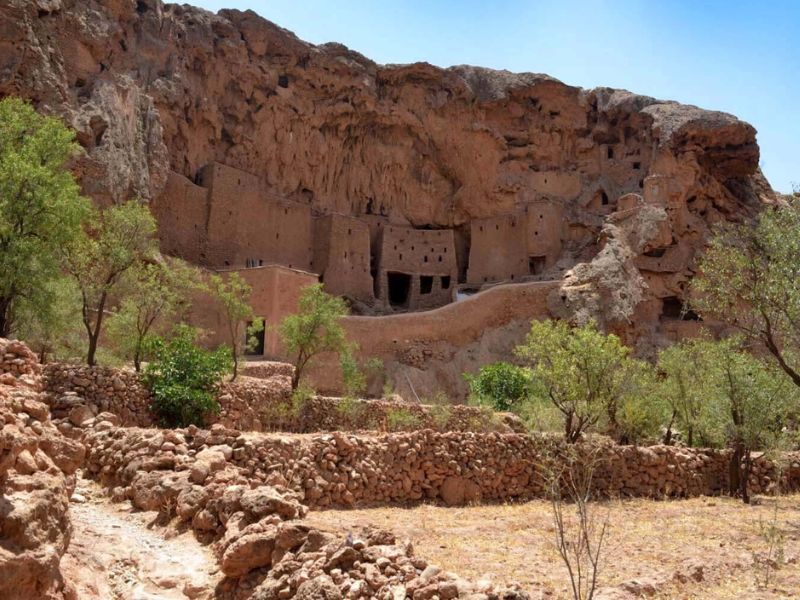 Ancient cliff dwellings built into a rocky hillside, surrounded by desert vegetation and stone pathways.