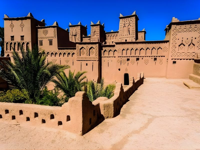 A view of an ancient adobe fortress with intricate geometric designs under a clear blue sky, surrounded by lush palm trees.