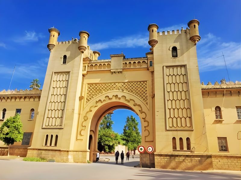 A grand archway with decorative towers and intricate designs, leading into a lush green area with people walking beneath it.
