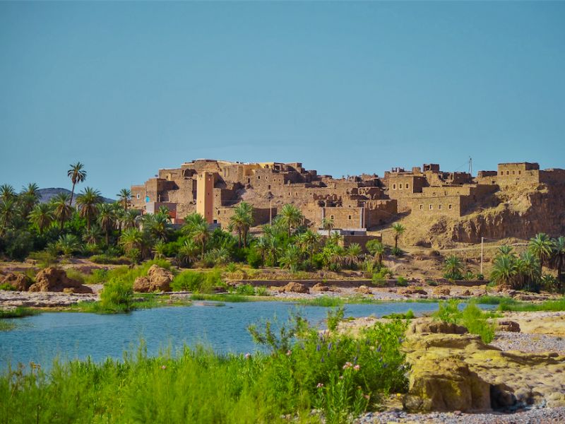 View of an ancient village on a hillside, surrounded by palm trees and a river in the foreground, under a clear blue sky.