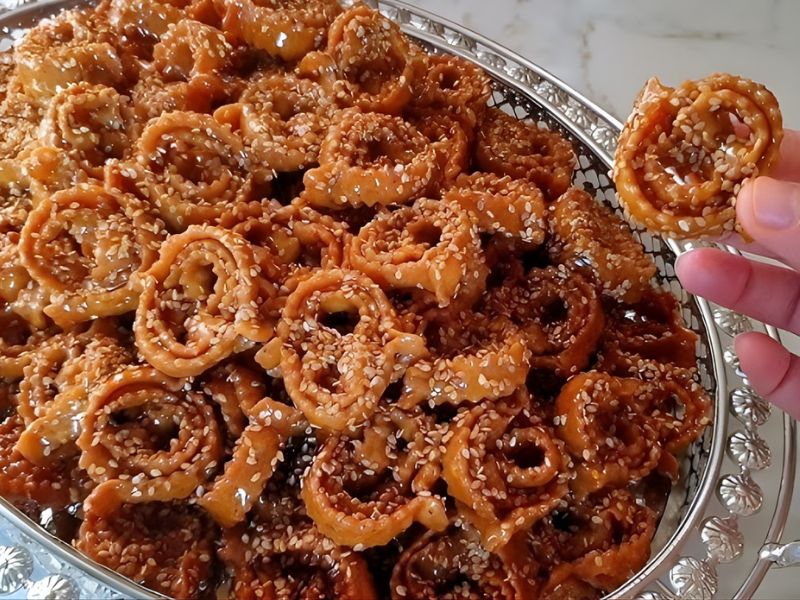 A close-up of a plate filled with sweet, sesame-coated circular pastries, with one pastry being held up by a hand.