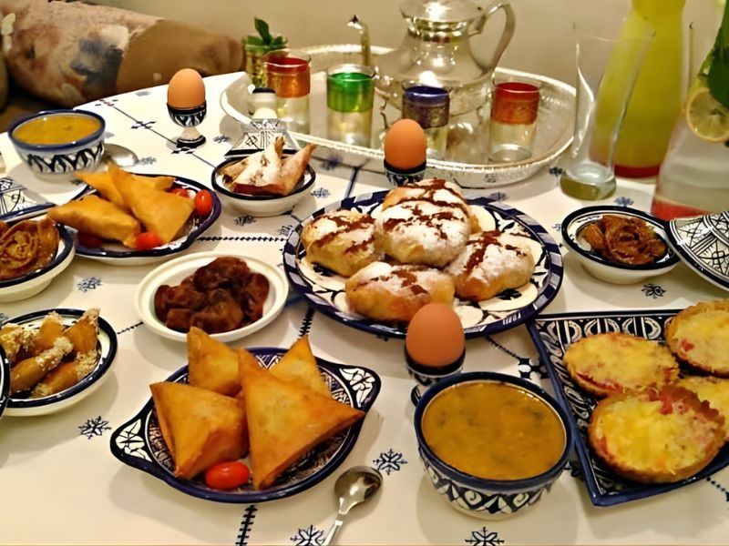 A beautifully arranged spread of assorted Moroccan dishes on a decorative tablecloth, featuring samosas, pastries, eggs, and bowls of soup, accompanied by colorful glasses and a traditional teapot.