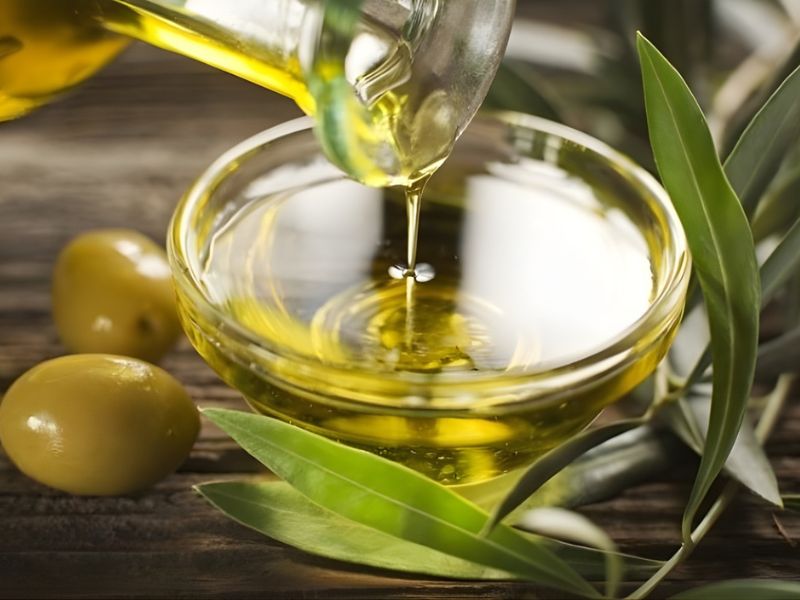 A glass bowl of olive oil being poured from a bottle, surrounded by green olives and olive leaves on a wooden surface.
